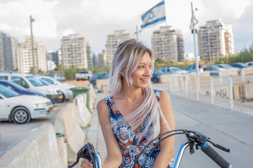 A smiling woman in a blue floral outfit beside a bicycle on the Ashdod waterfront.