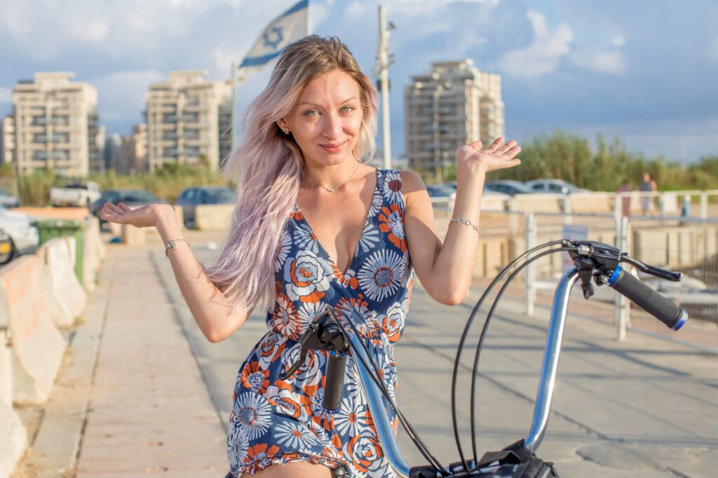 A woman in a blue floral outfit raising her hands playfully beside a bicycle.